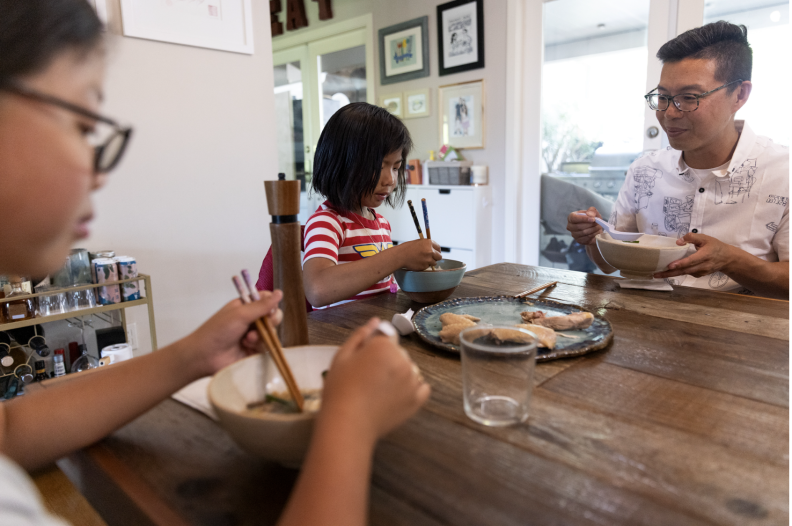 Three Asian people, two young girls and an older man, are sitting at a dining room table eating a bowl of broth, noodles, and onions. The girl in the foreground wears glasses and holds chopsticks and a spoon, the younger girl wears a red striped shirt and holds chopsticks, and the older man wears a white patterned shirt and smiles while holding his bowl and spoon. The window behind the table lets in white natural light, which illuminates the room and table.
