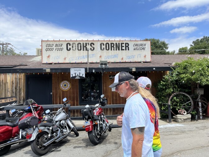A couple walk past a row of motorcycles lined up outside the a single-story building lined with vertical wood planks, holiday lights, and a faded sign that reads: "Cook's Corner, Cold Beer, Good Food, Children Welcome."