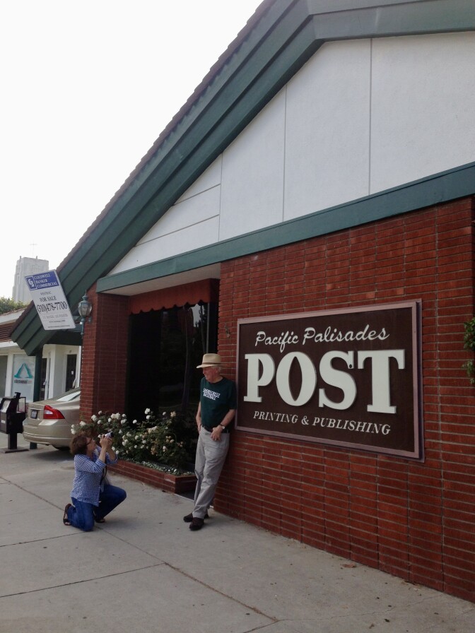 A man stands next to a sign that says "Pacific Palisades Post". He is in green and wears a hat and gray pants. A woman in a blue shirt and jeans kneels to take a picture of him.