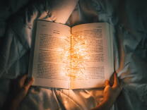 A book lies open on a bed's comforter, with two hands holding each corner at the bottom. It is dark and the only light is coming from a cluster of fairy lights that lie at the crease of the open book, illuminating the words. 
