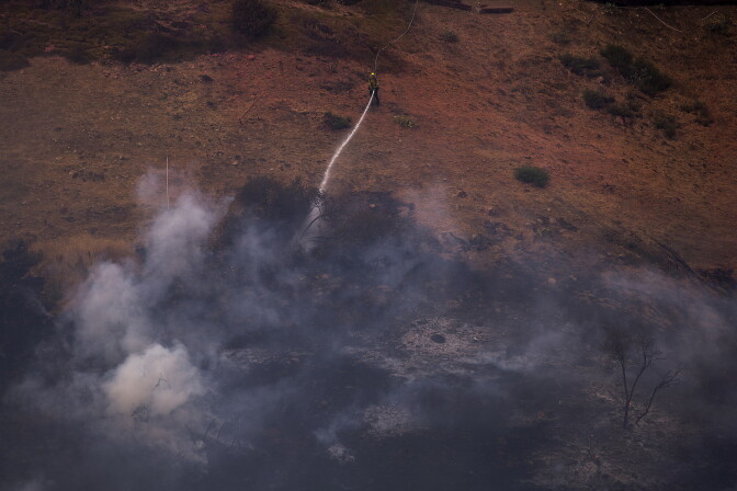 A firefighter, shown from above, sprays water onto a massive cloud of smoke coming up from the ground on a dry hillside. The air is hazy with smoke as well from what appears to be a fire that was just put out.