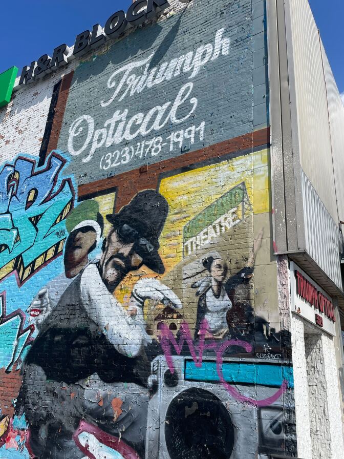 A colorful mural on the side of a building. The sign on the front of the building is difficult to see, but still visible. It reads "Triumph Optical" in red lettering. At the top of the wall is a black and green sign that reads "H&R Block." The mural on the brick wall below it features a man with sunglasses, a mustache and a fedora; another person with a baseball cap; and a woman wearing a black and white baseball tee and raising her arm. A painting of a boombox is visible on the bottom, blue and teal graffiti bubble lettering on the left, and a depiction of the "HIGHLAND THEATRE" sign in green and white lettering.