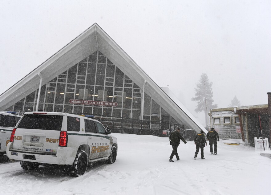 A white Sheriff's SUV sits in front of a building with a triangular style roof. Three people in dark clothing/jackets are walking in the snow to the right of the SUV