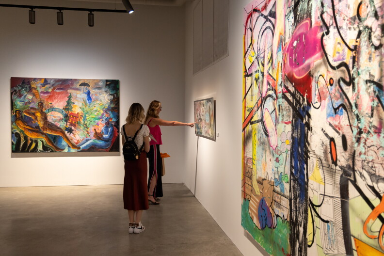 A woman stands looking at another, who points to a piece of art on the wall. There are two larger pieces hanging nearby. The piece in the background has multiple nude figures reclining, with one person wearing a blue suit.