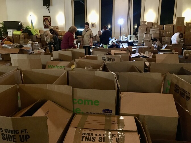 A room is filled with open cardboard boxes full of Ukraine relief supplies. Several people are in the back of the room filling boxes.