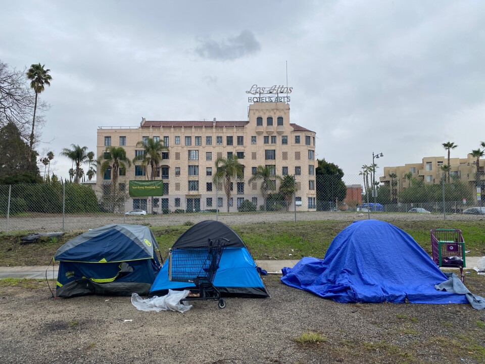 Three blue tents used by people experiencing homelessness are lined up on along a sidewalk. Behind them is a hotel.