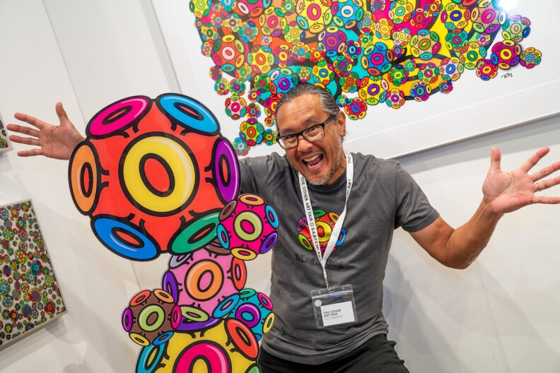 a man in a gray shirt smiles and holds out his arms around a multi-colored sculpture/work of art
