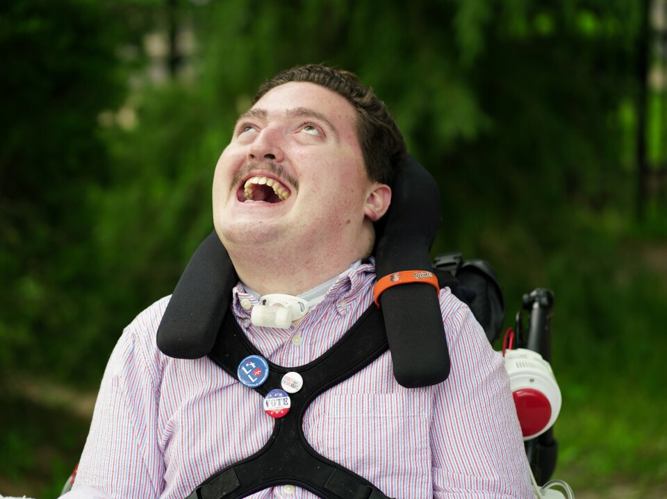 A man with light-toned skin who appears to be in his 30s sits in a wheelchair. His head is thrown back in a laugh. He has padding around his neck, a safety strap across his chest and what appears to be a tracheostomy tube. He wears a white and red pinstriped button-up shirt. 