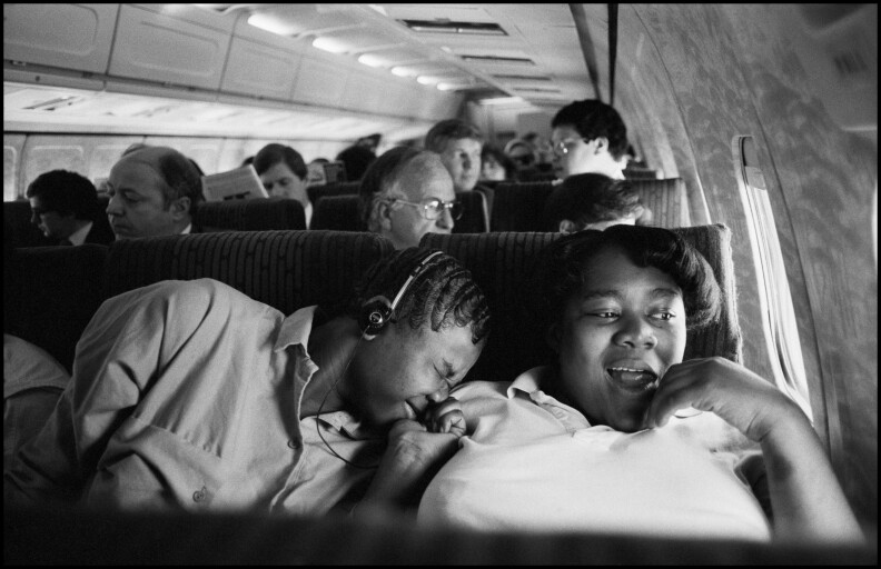 One person wears headphones and leans on the shoulder of a friend on a bus