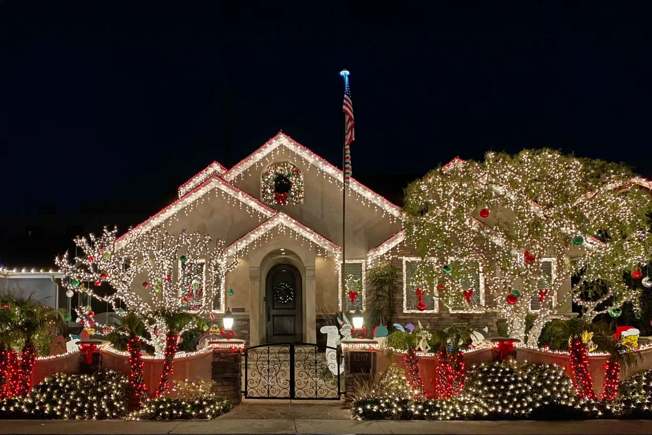 A home is decorated with white and red lights for the holidays. Two trees in front of the homes are also decorated with lights as are the bushes on the sidewalk.