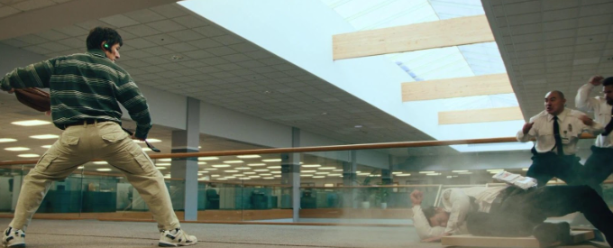 Ke Huy Quan in character as Alpha Waymon stands with his back to the camera and preps a fanny pack as a weapon. He is fighting three security guards on the right of the image.