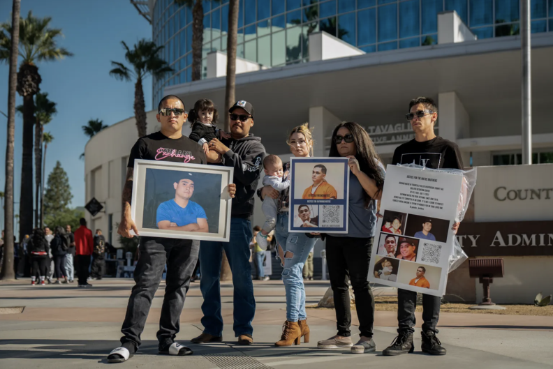 A group of three men and two women, all with brown skin tone, hold signs and pictures of a man they are memorializing. They are standing in front of what appears to be a government building. One of the men and one of the women are each holding a baby.