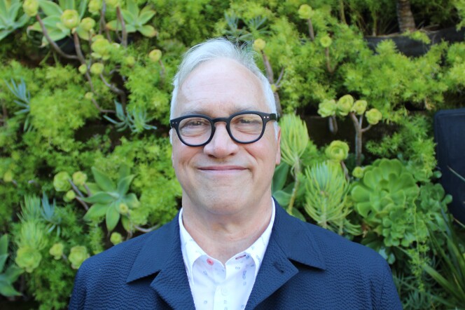 A man with white hair and glasses stands in front of greenery. 