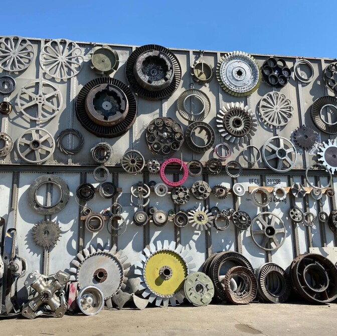 A outdoor wall with a variety of what look like different sizes of metal hubcaps and gears hung on it. Blue sky is visible over the wall.