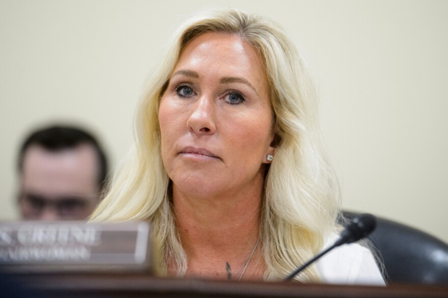 A woman with light-tone skin and blond hair sits in front of a mic and name card that's blurred.