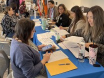 Seven people sit on both sides of a table draped by a blue tablecloth and covered with paperwork.
