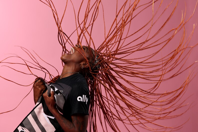 Messiah Bright, a Black woman, tosses her head back and yells while clutching her jersey, as if celebrating during a game. Her red-orange braids are captured mid-flip, fanned out around her. She's standing in from of a pink background.