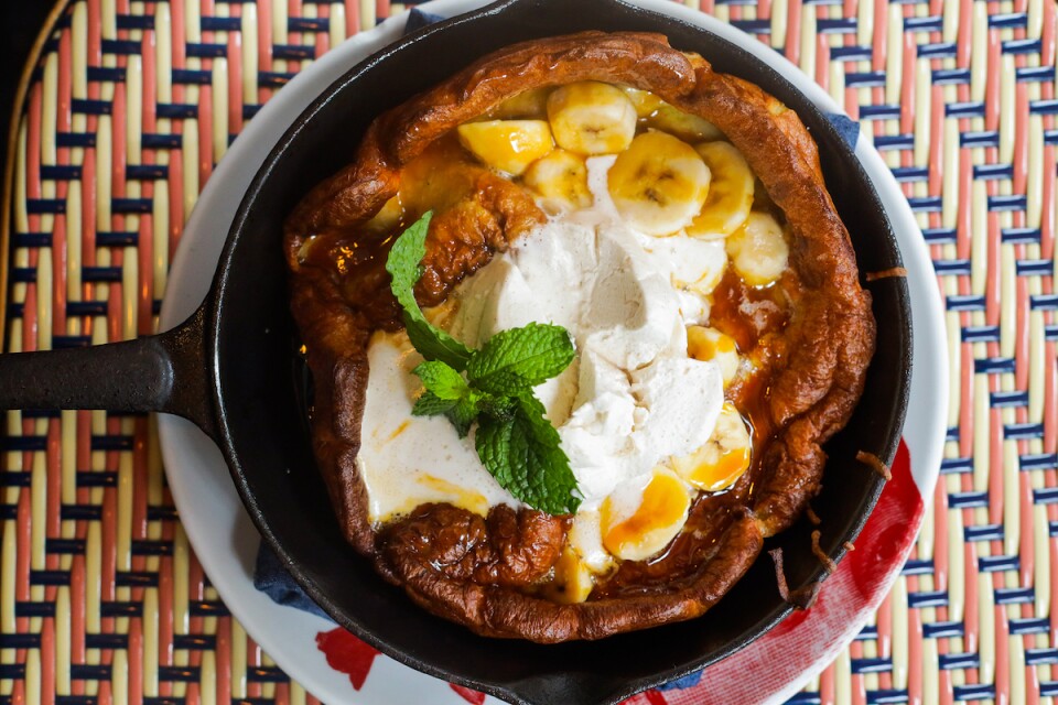 a skillet of a Dutch baby pancake with topped with bananas, whipped cream and a mint leaf.