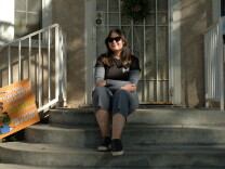 A young woman wearing a black t-shirt and grey pants sits on concrete steps leading to the front door of her house. 