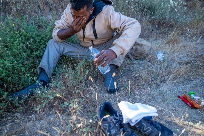 A man with a water bottle sits in some shrubs with a beige hoodie and grey jeans on 
