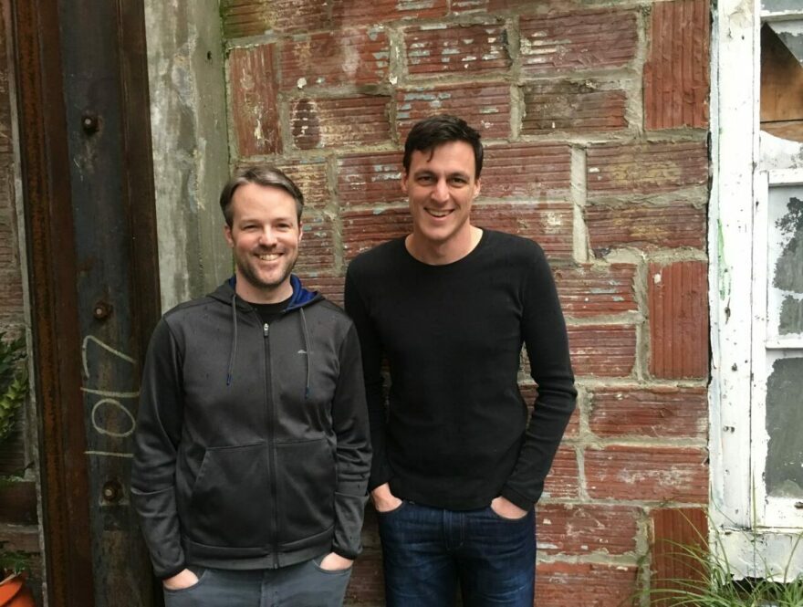 Two men with light tone skin have their hands in their pockets and smile for a photo in front of a red brick wall.
