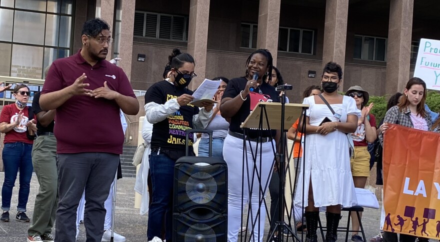 Amber Sam stands at a podium surrounded by a crowd of people who attended a rally to call on LA County to invest more in mental health treatment beds. Sam wears white pants and a black shirt.
