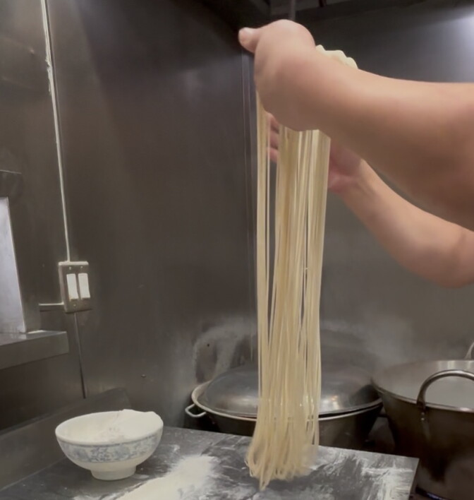 A cook’s hands, with light-to-medium skin, hold up a long bundle of freshly pulled noodles over a floured steel counter. A ball of dough and a bowl of flour sit nearby, with large pots on the stove in the background.