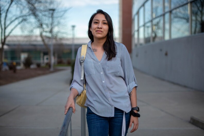 A young woman with light Brown skin and straight dark hair stands on a set of concrete steps, leaning against a steel railing. She wears a loose-fitting blue-and-white striped button-down shirt, jeans and a yellow backpack on one shoulder.