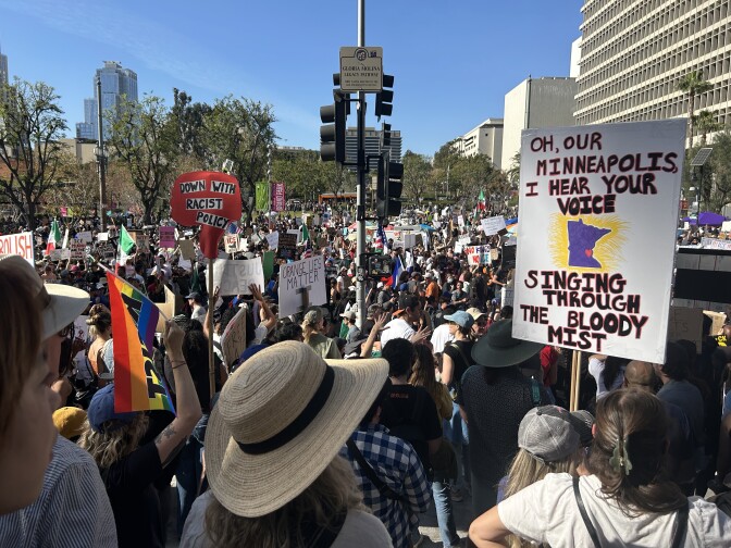 A large crowd of people gathered in downtown Los Angeles is photographed from behind. Some of them are holding signs. One reads, "Oh, our Minneapolis, I hear your voice. Singing through the bloody mist."