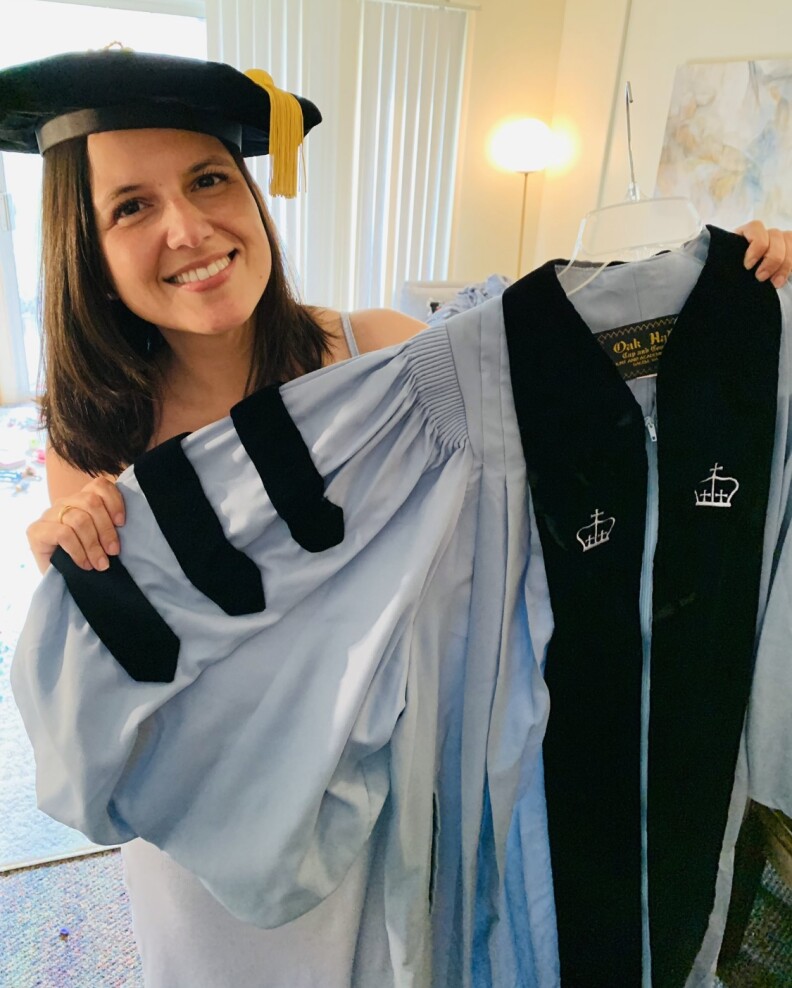 A smiling woman with shoulder-length hair wears a commencement tam and hold up a light blue and black gown. 