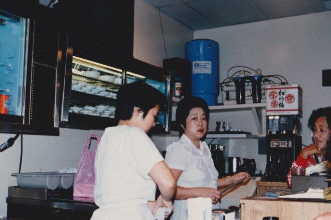 A photo of two Asian women — one is looking at the camera while the other has her back to the lens —  in white working at a restaurant counter, talking to another Asian woman 