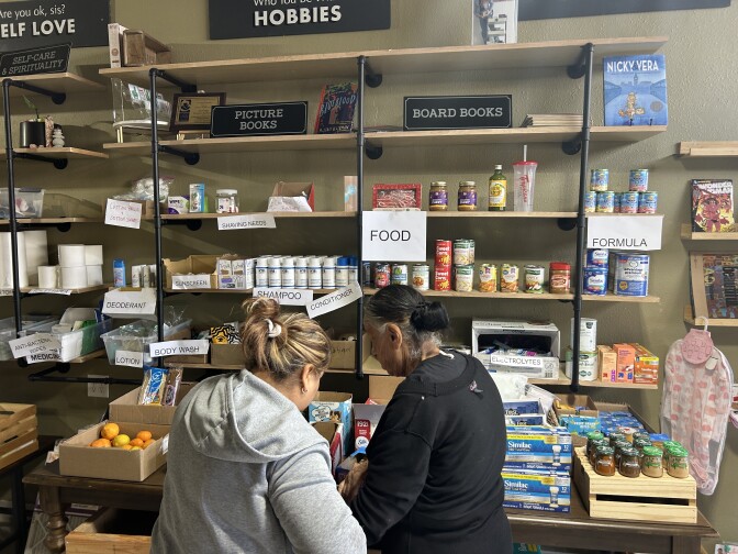 Two people stand in front of a table and shelves laden with household and personal goods, including baby formula, shampoo and lotions. 