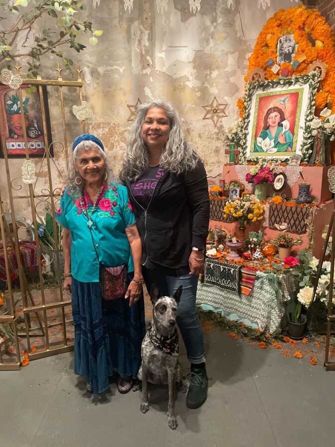 Two women with gray wavy hair stand with a dog in front of an ofrenda, or altar, covered in flowers, paintings and photos to remember loved one who died. 