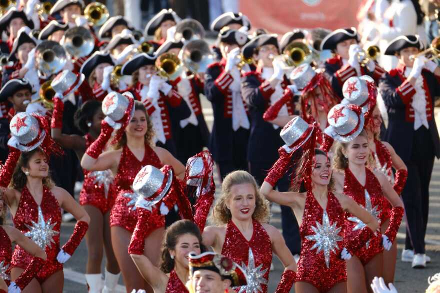 Women in sparkling red outfits and glittering silver top hats march in a parade ahead of a band dressed in colonial American soldier uniforms.