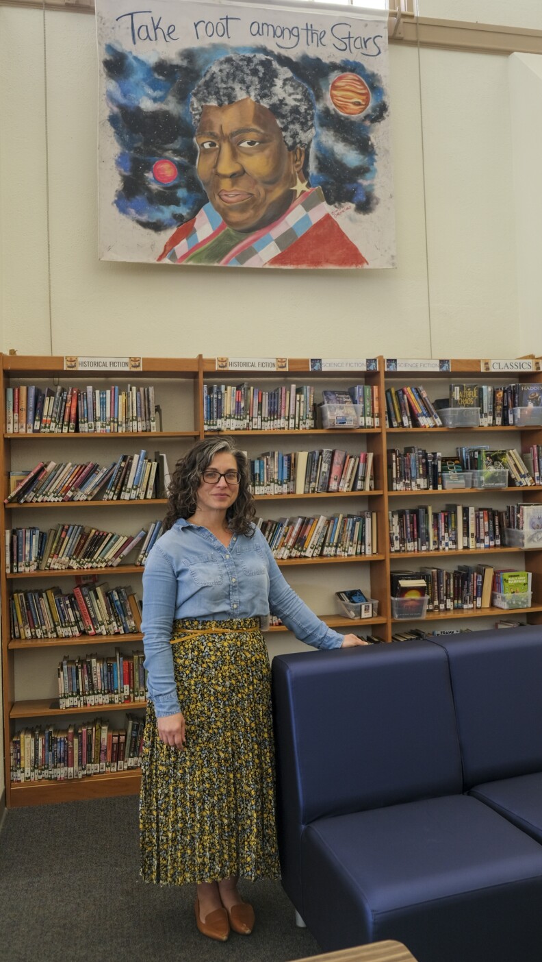 A woman with light skin tone, shoulder-length curly hair and glasses stands in a room filled with books. There is a chalk mural overhead of a woman with dark skin tone and short curly hair surrounded by space.