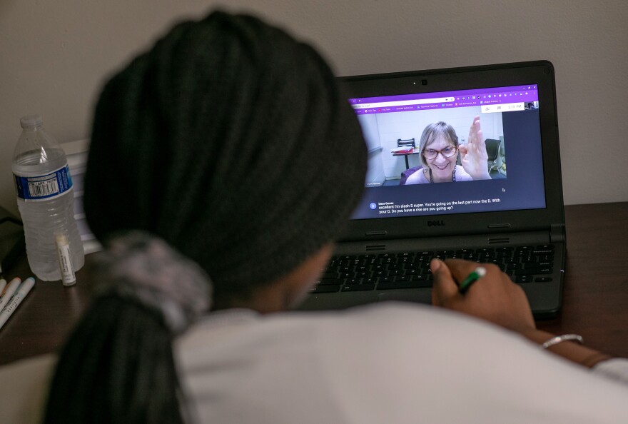 The camera looks over the shoulder of a student who is watching a teacher on her computer during an online class.