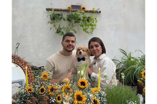 A couple sits at a table surrounded with sunflowers between a dog. 