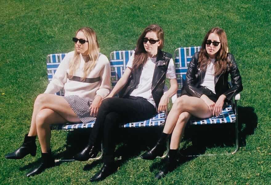 The three members of Haim sit on three plastic lawnchairs in sunglasses as they look to the right.