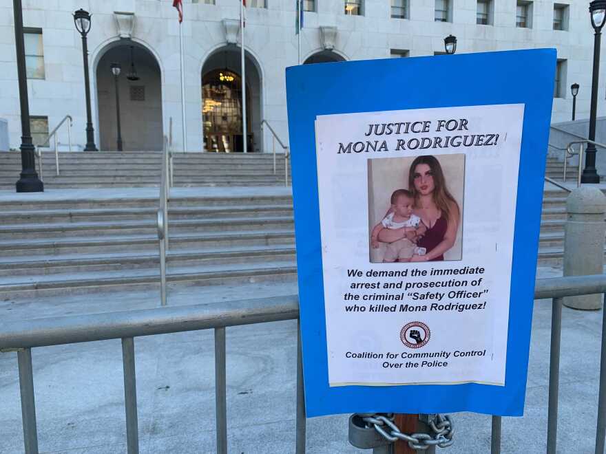 A small poster with a blue border is affixed to a railing in front of a building, with two sets of steps going up behind it. On a white background, the poster reads: "Justice for Mona Rodriguez!" at the top. Underneath those words is a color photo of Rodriguez holding her infant son. Below the photo is says: "We demand the immediate arrest and prosecution of the criminal 'Safety Officer' who killed Mona Rodriguez!" It is signed at the bottom by the Coalition for Community Control Over the Police.
