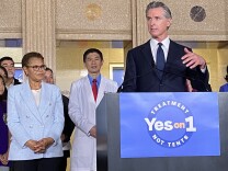  Gov. Gavin Newsom wears a blue suit and stands at a podium. The podium has a blue poster on it that reads: "yes on 1: treatment not tents" To his right, Newsom is joined by LA Mayor Karen Bass, who has her hands folded in front of her. 