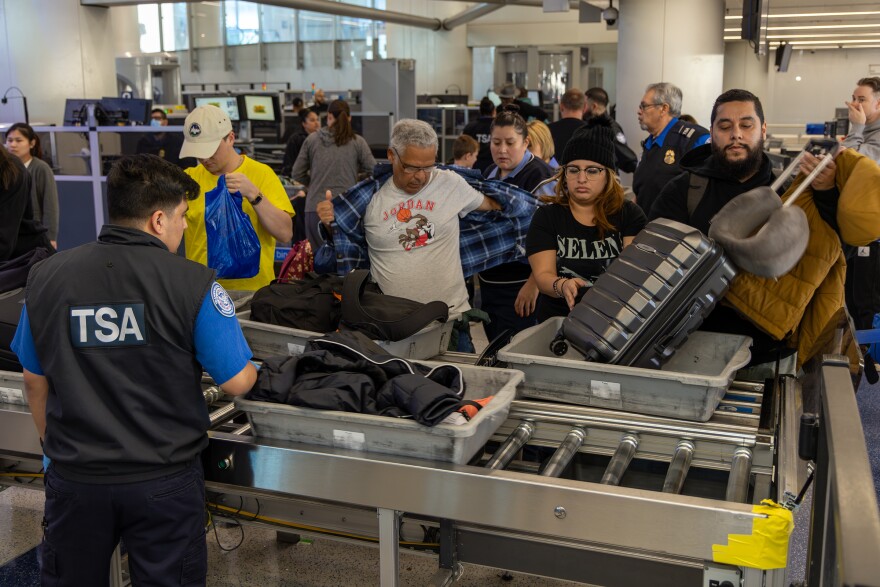 A wide look at a group of men and women with different skin tones as they load personal belongings, like suitcases and jackets, into gray bins on a conveyor belt for TSA screening.