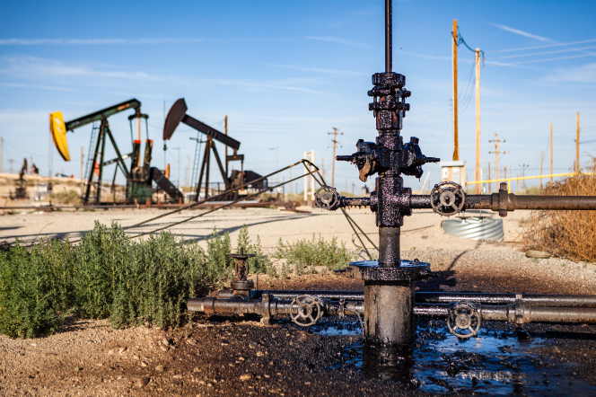 Several oil derricks and a pipeline surrounded by a pool of oil are shown in a dusty field