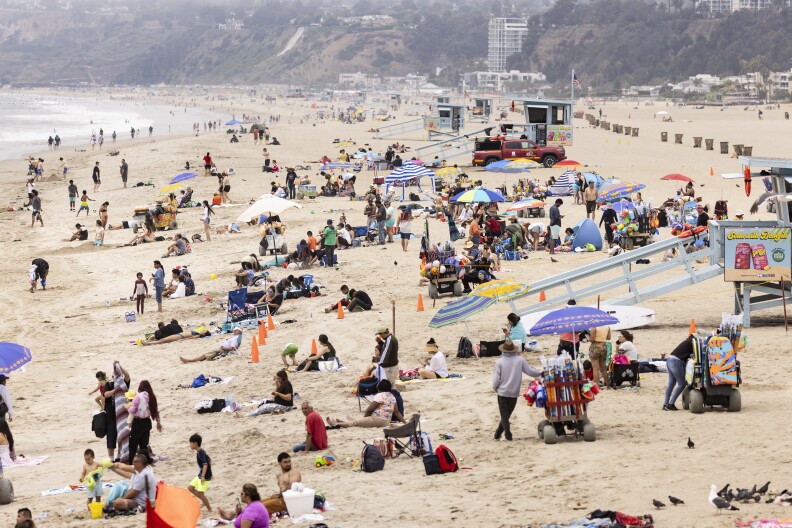 A sandy beach packed full of people, umbrellas, and towels. Several light blue lifeguard stations are scattered along the coast, and the ocean waters can be seen in the distance.