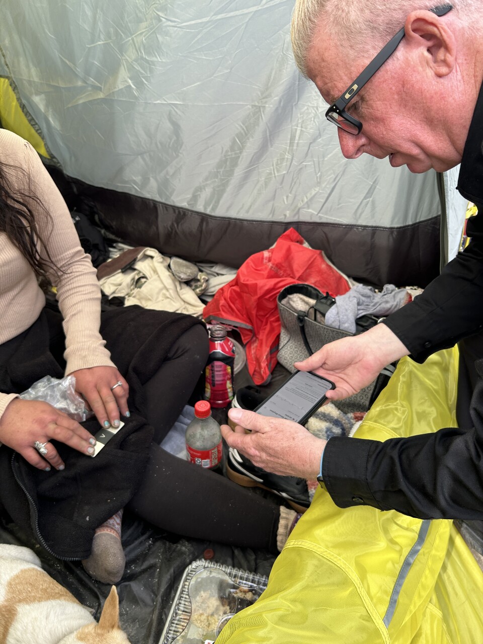The inside of a tent showing, on the left side, a woman's hands with her fingers pressed on a mobile EKG monitor, and on the right, a man's face in profile. He's holding a cell phone monitoring the device. On the floor of the tent are soda bottles and clothes, and you can part of a small white and tan dog. 