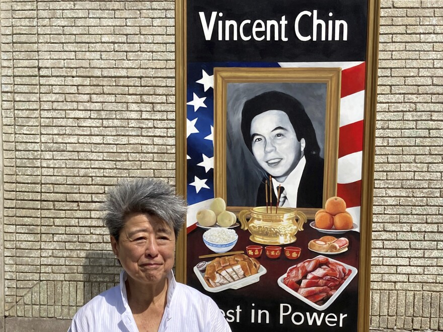 Helen Zia has shock of gray hair against a tan brick wall with a portrait of Vincent Chin