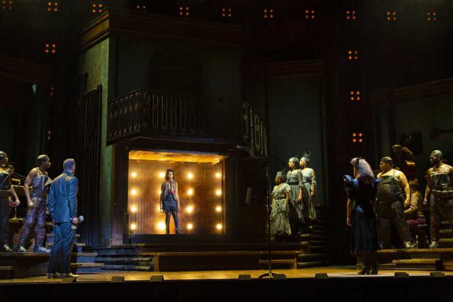 Several people on a dark stage, looking toward a woman standing in a lit box center stage.