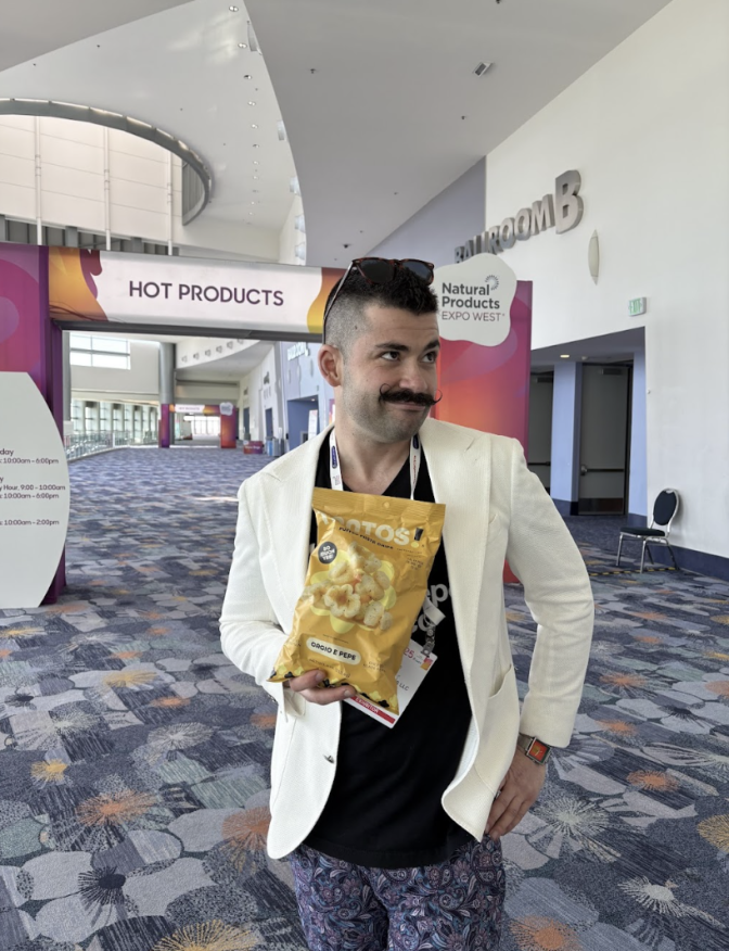 A medium skinner man with sunglasses perched on his head and a black mustache, wearing a white jacket, is posing with a large packet of chips in his hands, 