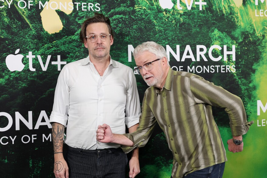 Two people stand in front of a backdrop that reads "Monarch Legacy of Monsters" on it.