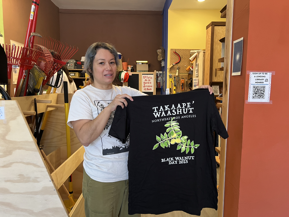Executive Director Brenda Contreras poses with one of the many t-shirts Coyotl+Macehualli has made and has on site at the lending library. This shirt design was done by local Indigenous illustrator Samantha Morales Johnson (Tongva)