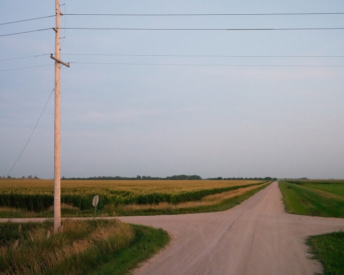 A rural dirt crossroads has a single stop sign and a telephone pole at the near left corner.